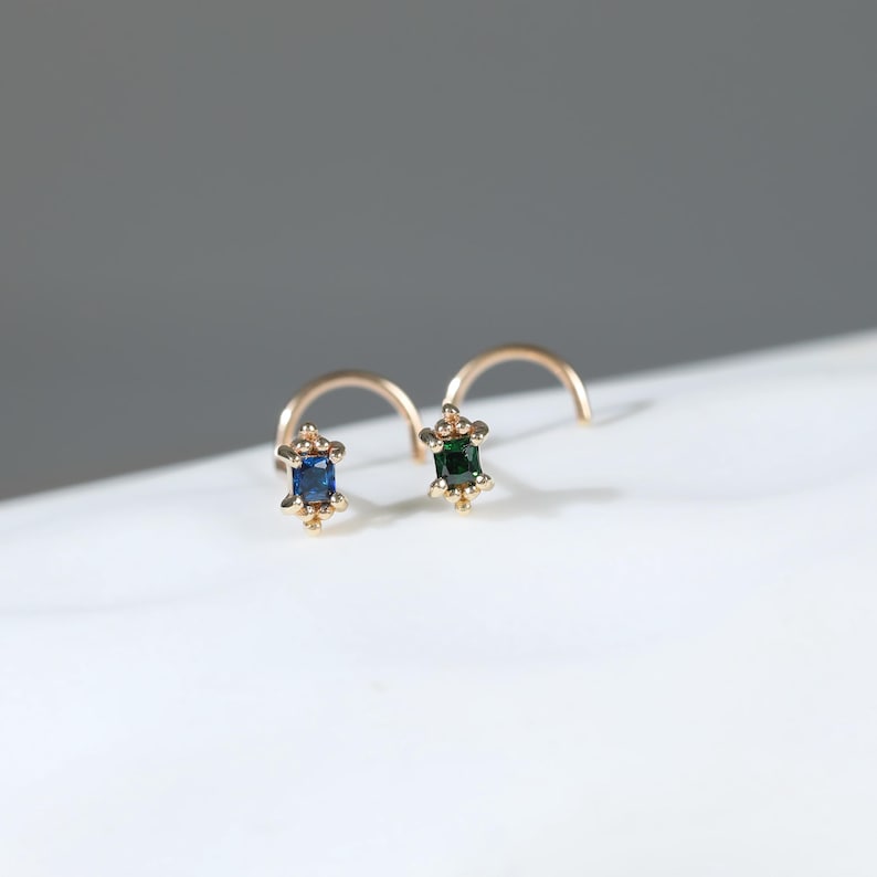 May include: Two gold nose rings featuring small rectangular gemstones. One ring holds a deep blue sapphire-coloured stone, while the other holds a dark green emerald-coloured stone. Each setting is accented with tiny gold beads at the corners.