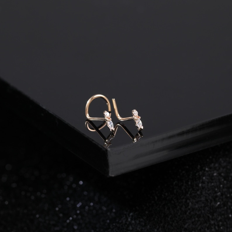 Peut inclure: Un bijou de nez en or rose avec une tige courb&eacute;e en forme de L et un motif d&eacute;licat orn&eacute; de deux petites pierres transparentes taille marquise. Le m&eacute;tal pr&eacute;sente une finition chaude et les pierres sont dispos&eacute;es en ligne sur la tige.