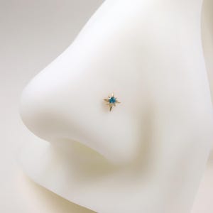 May include: A gold-colored nose stud featuring a starburst design with a central teal-colored gemstone. The jewelry is set against a white background, highlighting the delicate details of the piercing.