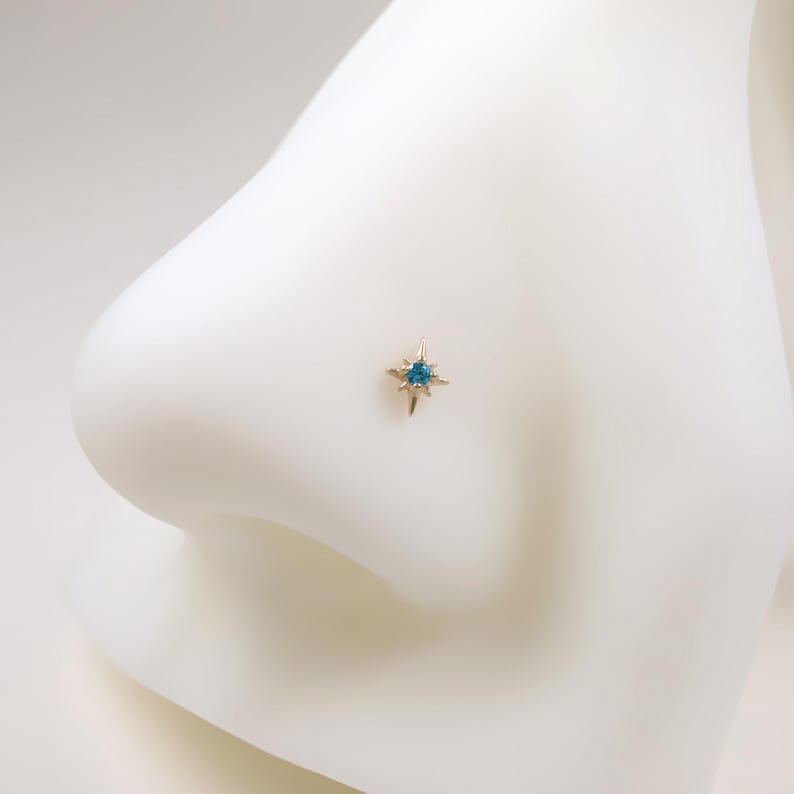 May include: A gold-colored nose stud featuring a starburst design with a central teal-colored gemstone. The jewelry is set against a white background, highlighting the delicate details of the piercing.