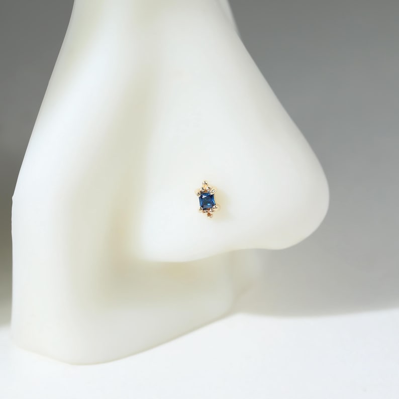 May include: A gold nose stud featuring a rectangular, deep blue gemstone set in a prong setting. The small, elegant piece of jewelry is positioned on a white, abstract nose-shaped display form, highlighting the contrast between the gold and blue.