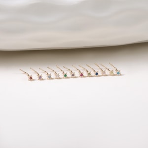 May include: A collection of gold-colored nose studs, each featuring a star-shaped design with a central gemstone. The gemstones are in various colors, including red, purple, blue, and green. The studs are arranged in a row on a white surface.