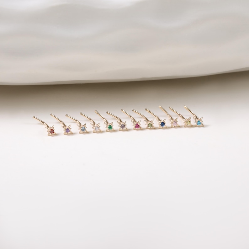 May include: A collection of gold-colored nose studs, each featuring a star-shaped design with a central gemstone. The gemstones are in various colors, including red, purple, blue, and green. The studs are arranged in a row on a white surface.