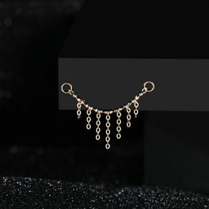 May include: A gold-toned metal jewelry connector featuring a curved horizontal chain with seven dangling vertical chain segments of varying lengths, finished with a small circular jump ring at each end for attachment purposes.