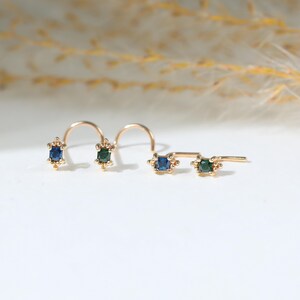 May include: Four gold nose studs featuring small rectangular gemstones. Two studs have blue stones and two have green stones. Each setting is accented with tiny gold beads at the top and bottom, attached to curved or L-shaped gold posts.