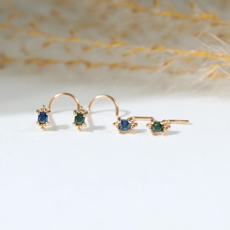 May include: Four gold nose studs featuring small rectangular gemstones. Two studs have blue stones and two have green stones. Each setting is accented with tiny gold beads at the top and bottom, attached to curved or L-shaped gold posts.