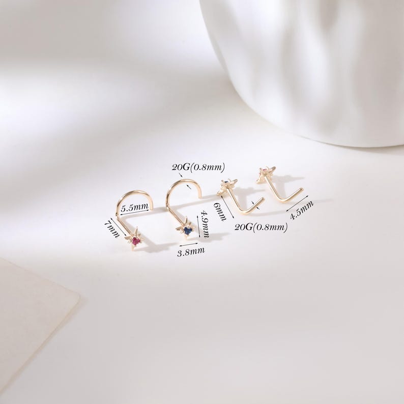 May include: Four gold-colored nose studs with star-shaped designs and gemstone accents. The studs vary in size, with measurements in millimeters and gauge (20G/0.8mm). One stud has a red gemstone, another a blue gemstone, and the others have clear gemstones.