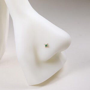 May include: A gold nose stud featuring a starburst design with a central emerald-green gemstone. The jewelry is set on a white mannequin nose, showcasing the stud's delicate details and color contrast.