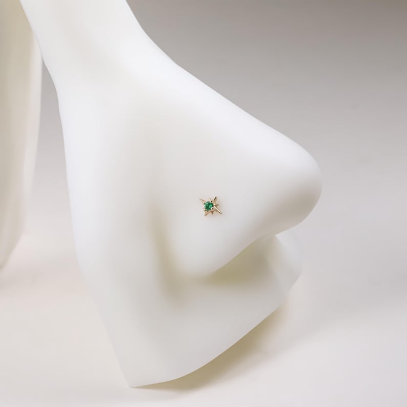 May include: A gold nose stud featuring a starburst design with a central emerald-green gemstone. The jewelry is set on a white mannequin nose, showcasing the stud's delicate details and color contrast.