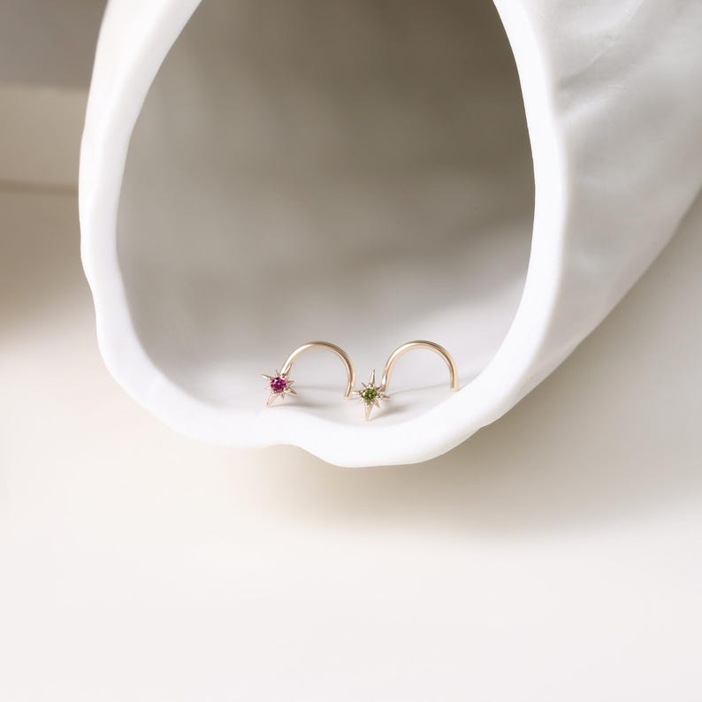 May include: Two gold nose rings, each featuring a star-shaped design. One star has a red gemstone, and the other has a green gemstone. The rings are displayed on a white, curved surface.