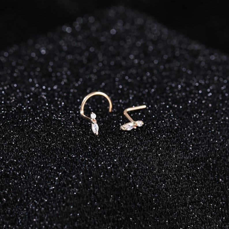 Peut inclure: Deux bijoux de nez dor&eacute;s dot&eacute;s d'une tige courb&eacute;e et d'une extr&eacute;mit&eacute; d&eacute;corative orn&eacute;e d'une petite pierre ronde transparente et d'une pierre transparente taille marquise. Les bijoux sont dispos&eacute;s c&ocirc;te &agrave; c&ocirc;te, r&eacute;v&eacute;lant le m&eacute;tal d&eacute;licat.