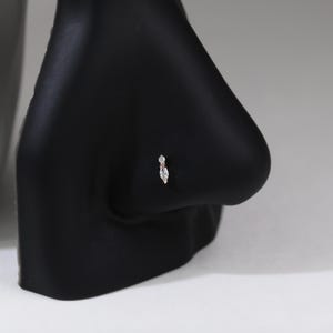 Peut inclure: Un bijou de nez en or compos&eacute; d'une petite pierre transparente ronde plac&eacute;e au-dessus d'une pierre transparente plus grande en taille marquise. Le bijou est pr&eacute;sent&eacute; sur un support de mannequin noir en forme de nez, montrant les deux pierres.