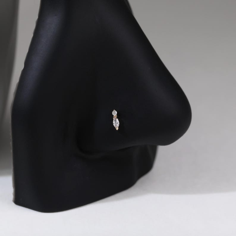 Peut inclure: Un bijou de nez en or compos&eacute; d'une petite pierre transparente ronde plac&eacute;e au-dessus d'une pierre transparente plus grande en taille marquise. Le bijou est pr&eacute;sent&eacute; sur un support de mannequin noir en forme de nez, montrant les deux pierres.