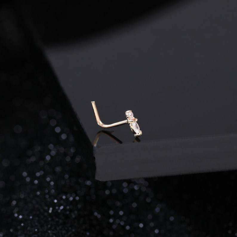Peut inclure: Un bijou de nez en forme de L en m&eacute;tal dor&eacute;, dot&eacute; d'une t&ecirc;te d&eacute;corative orn&eacute;e d'une petite pierre ronde transparente et d'une pierre transparente taille marquise dispos&eacute;es en ligne, offrant une apparence d&eacute;licate et raffin&eacute;e pour le visage.
