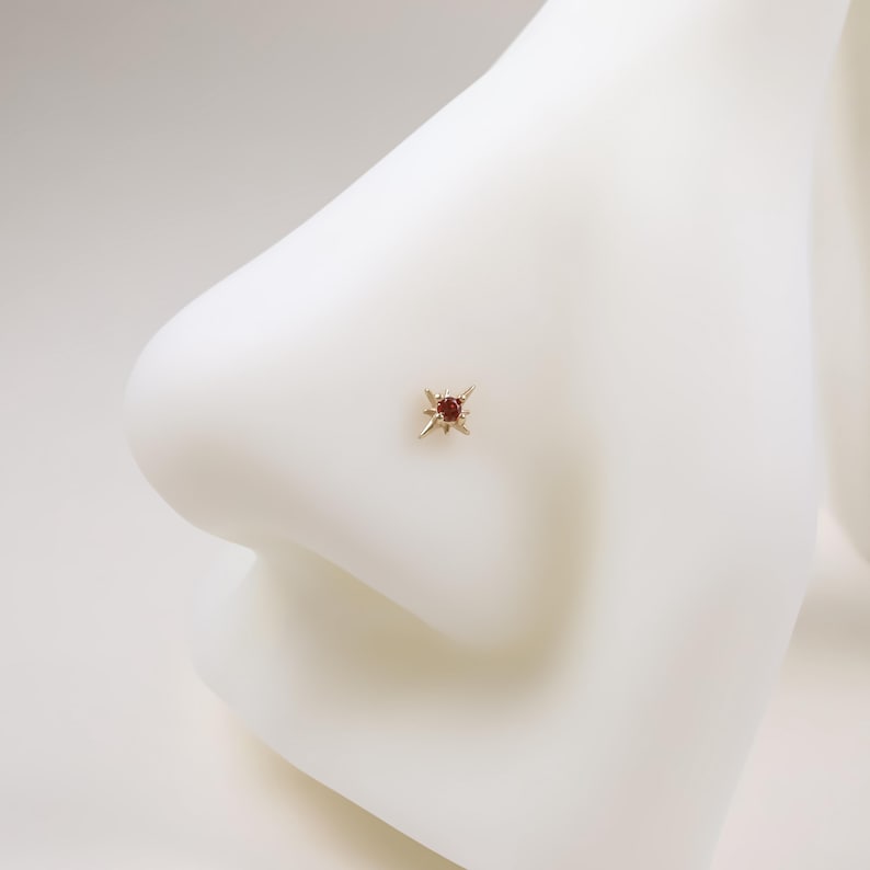 May include: A gold-colored nose stud featuring a starburst design with a central, round, red gemstone. The jewelry is set against a white background, highlighting the stud's delicate details and color.