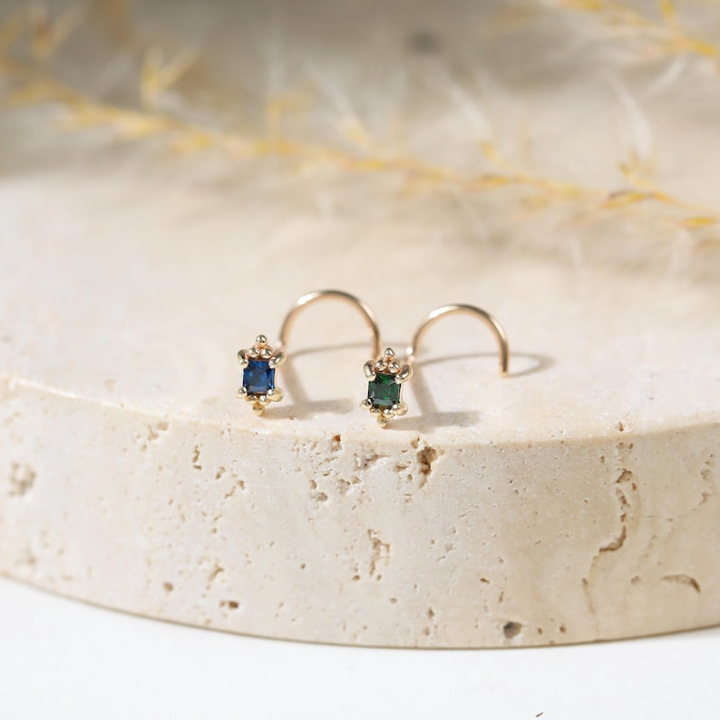 May include: Two gold nose rings featuring small rectangular gemstones. One nose ring has a deep blue rectangular stone, and the other has a dark green rectangular stone. Both pieces are set in gold metal with small decorative beads at the top and bottom.