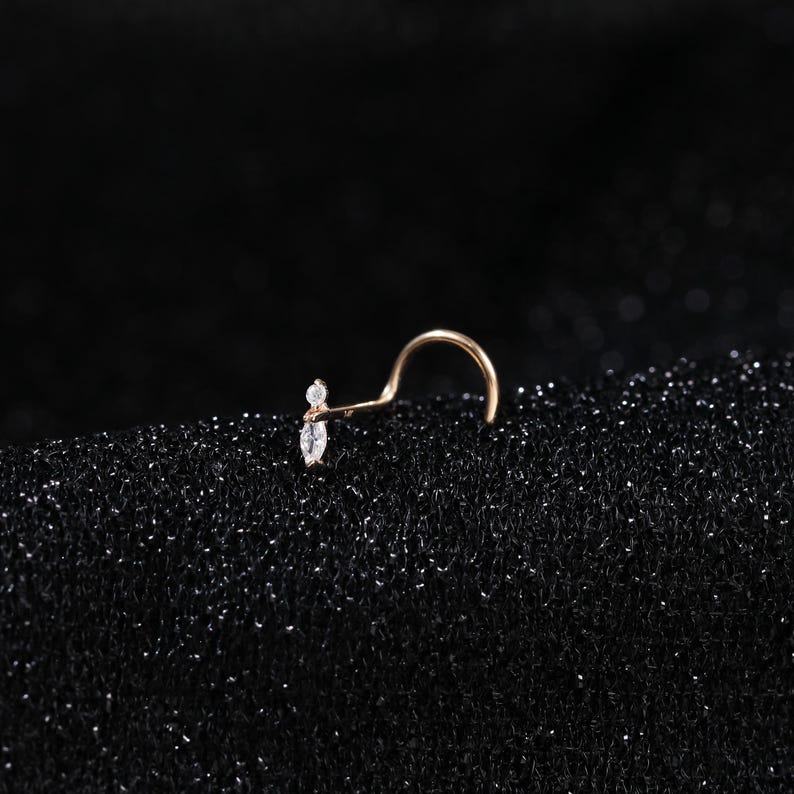 Peut inclure: Un piercing de nez en or rose avec une tige courb&eacute;e et une partie d&eacute;corative compos&eacute;e d'une petite pierre ronde transparente et d'une pierre transparente en forme de marquise dispos&eacute;es verticalement. Le m&eacute;tal a une finition chaude.