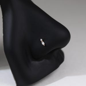 Peut inclure: Un piercing de nez compos&eacute; d'une petite pierre transparente ronde surmontant une pierre transparente plus grande en forme de marquise, le tout serti dans une monture m&eacute;tallique. Le bijou est pr&eacute;sent&eacute; sur un mannequin noir mat.