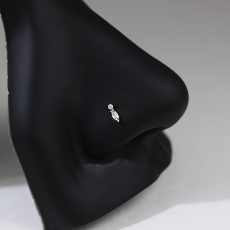 Peut inclure: Un piercing de nez compos&eacute; d'une petite pierre transparente ronde surmontant une pierre transparente plus grande en forme de marquise, le tout serti dans une monture m&eacute;tallique. Le bijou est pr&eacute;sent&eacute; sur un mannequin noir mat.