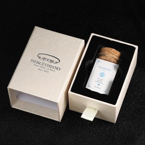 May include: A beige textured sliding gift box with the text PIERCETHEORY Every Stone, A Story since 2025. Inside, a glass vial with a cork stopper contains a label reading Aquamarine, Clear, calm, and full of beautiful dreams, Pierce Theory.