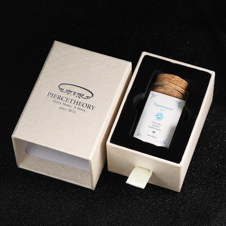 May include: A beige textured sliding gift box with the text PIERCETHEORY Every Stone, A Story since 2025. Inside, a glass vial with a cork stopper contains a label reading Aquamarine, Clear, calm, and full of beautiful dreams, Pierce Theory.