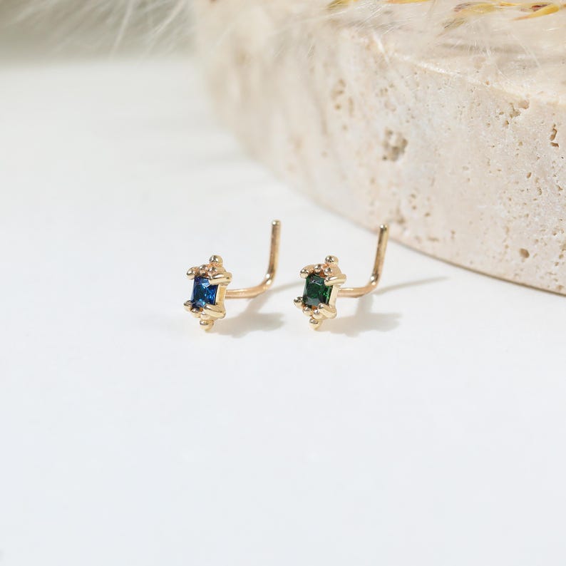 May include: Two L-shaped nose studs crafted from gold-toned metal. One stud features a rectangular blue gemstone, while the other features a rectangular green gemstone. Both studs are accented with small metal beads at the top and bottom edges.