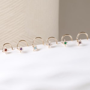 May include: A collection of gold-colored nose rings, each featuring a star-shaped gemstone in various colors. The gemstones include red, purple, blue, clear, green, and pink. The nose rings are displayed on a white surface.
