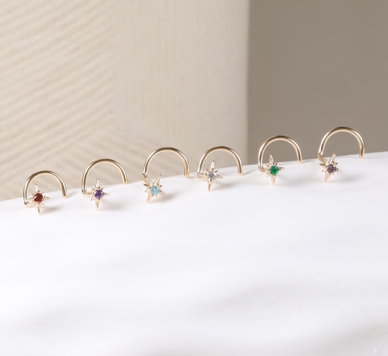 May include: A collection of gold-colored nose rings, each featuring a star-shaped gemstone in various colors. The gemstones include red, purple, blue, clear, green, and pink. The nose rings are displayed on a white surface.