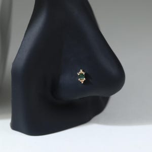 May include: A gold nose stud featuring a rectangular emerald green gemstone set in a vertical orientation. The setting includes small gold bead accents at the top and bottom of the stone, creating a decorative, symmetrical design on the metal post.