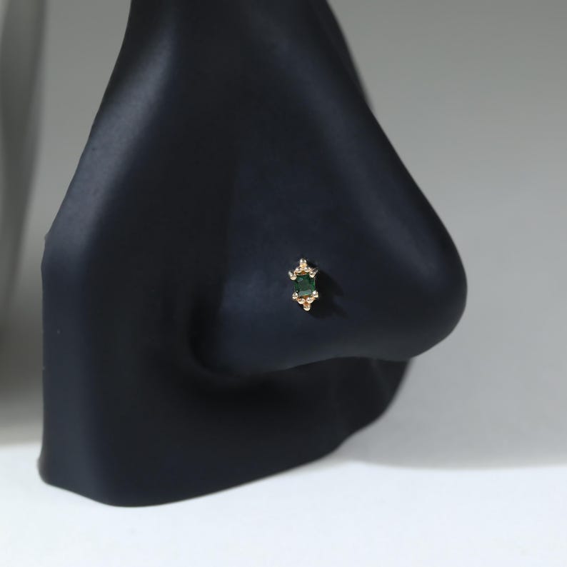 May include: A gold nose stud featuring a rectangular emerald green gemstone set in a vertical orientation. The setting includes small gold bead accents at the top and bottom of the stone, creating a decorative, symmetrical design on the metal post.
