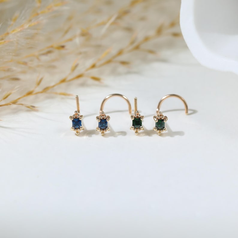 May include: Four gold nose studs arranged in a row. Each stud features a small rectangular gemstone in shades of blue and green, held by four tiny prongs. The metal posts are curved, designed for piercing, with a polished gold finish throughout.