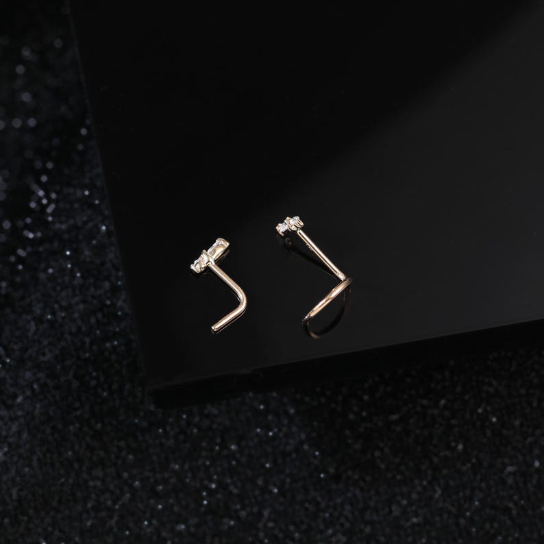 Peut inclure: Deux bijoux de nez en forme de L fabriqu&eacute;s dans un mat&eacute;riau m&eacute;tallique, chacun dot&eacute; d'une petite pierre pr&eacute;cieuse transparente de taille rectangulaire fix&eacute;e sur le dessus. Les bijoux sont plac&eacute;s c&ocirc;te &agrave; c&ocirc;te, montrant leur tige coud&eacute;e.