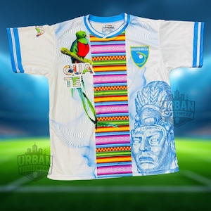May include: White football shirt with blue trim, showcasing a vibrant Guatemalan design. The shirt features a green and red bird illustration, the word "GUATE", and a blue emblem. The jersey also has a blue and white Aztec-style graphic.