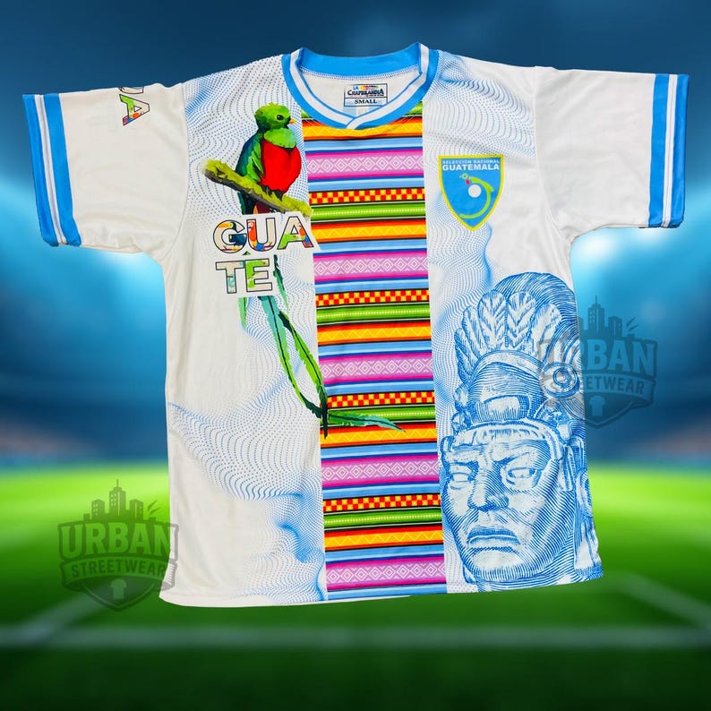 May include: White football shirt with blue trim, showcasing a vibrant Guatemalan design. The shirt features a green and red bird illustration, the word "GUATE", and a blue emblem. The jersey also has a blue and white Aztec-style graphic.