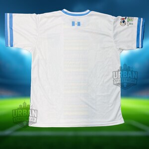 May include: White football shirt with light blue trim on the collar and sleeves. Includes a Guatemalan flag emblem and a sleeve logo. The shirt displays the "Urban Streetwear" logo.