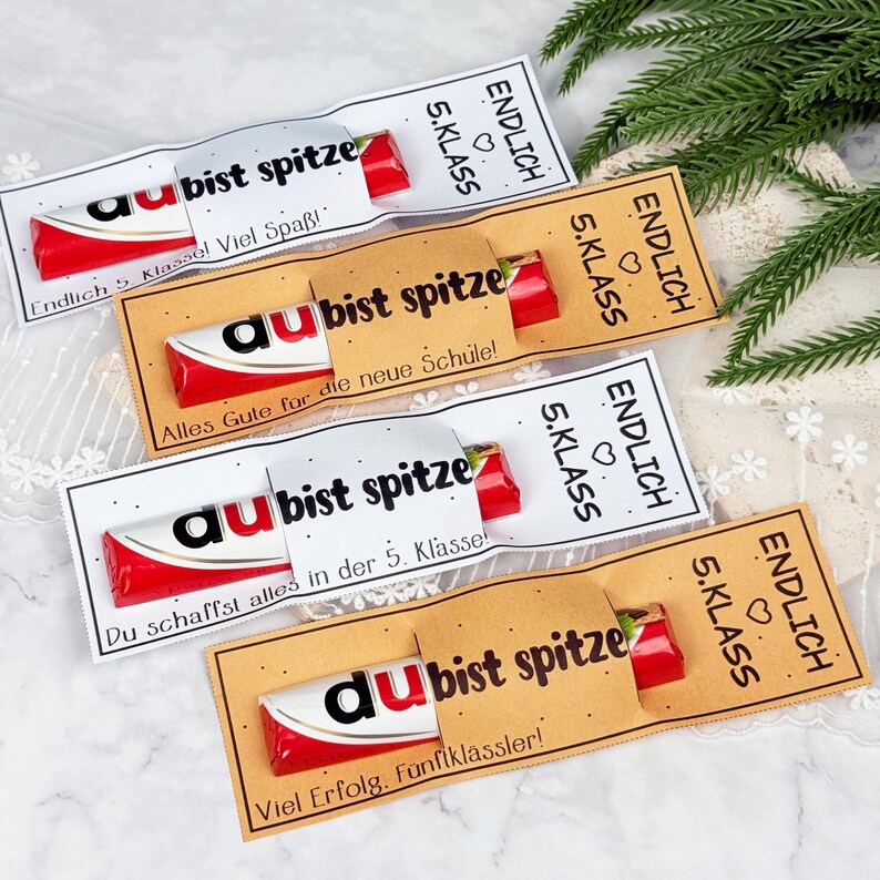 May include: Four rectangular paper sleeves holding red and white candy bars. The sleeves are white or tan, printed with black text including 'ENDLICH 5. KLASS', 'du bist spitze', and various German phrases. The design features small black dots.