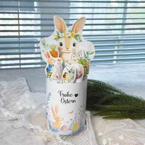 May include: A white cylindrical container with a bunny and floral design, holding Easter-themed cutouts. The container has the words "Frohe Ostern" and a heart. The cutouts feature bunnies, carrots, and eggs.