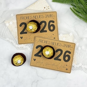 May include: Two kraft paper cards with the text "FROHES NEUES JAHR 2026" and a gold-wrapped chocolate candy. The cards also have the words "Glück, Gesundheit, Liebe" printed on them. A third candy is visible.