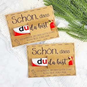 以下が含まれることがあります： 「Schön, dass du da bist」と「Ich wünsche dir einen guten Start in das neue Schuljahr」のテキストが入ったクラフト紙のカードが2枚。各カードには赤と白のチョコレートバーが入っています。左側には鉛筆のイラストがあります。