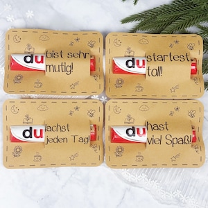 May include: Four rectangular kraft paper cards, each holding a red and white Duplo chocolate bar. The cards feature black line drawings of a moon, bee, flower, and cup, with German text: du bist sehr mutig, du startest toll, du lachst jeden Tag, du hast viel Spaß.