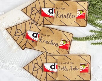 Puede incluir: Tarjetas de regalo de papel kraft con diseños de flechas, cada una con una barra de chocolate Kinder. Las tarjetas tienen el texto "bist der Knaller", "lasst es Krachen" y "as wird ein Tolls Fahr". El fondo es una superficie blanca con vegetación.