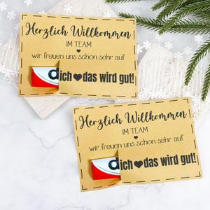 Puede incluir: Dos tarjetas de papel kraft con el texto "Herzlich Willkommen" y "dich das wird gut!" en escritura negra. Cada tarjeta tiene una pequeña barra de chocolate que asoma por una ranura. Las tarjetas están sobre una superficie blanca.