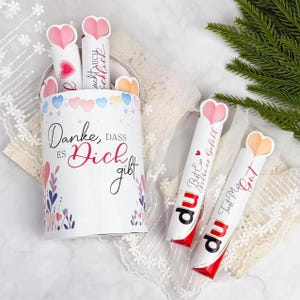 Peut inclure: Un récipient cylindrique blanc avec un couvercle, décoré de cœurs et de motifs floraux, contient plusieurs tubes en papier surmontés de cœurs. Le récipient porte l'inscription "Danke, dass es Dich gibt." Plusieurs tubes sont à l'extérieur.