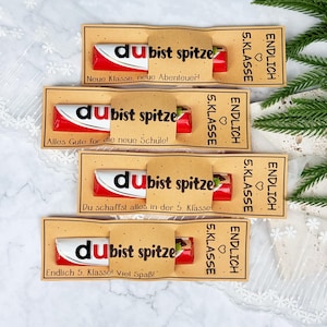 May include: Four chocolate bars wrapped in tan paper sleeves with black text. Each sleeve features the German phrase 'du bist spitze' and 'ENDLICH 5. KLASSE' with a heart icon. The individual messages include 'Neue Klasse, neue Abenteuer!', 'Alles Gute f&uuml;r die neue Schule!', 'Du schaffst alles in der 5. Klasse!', and 'Endlich 5. Klasse! Viel Spa&szlig;!'.