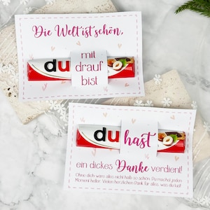 May include: Two rectangular white cards featuring pink heart patterns and German text, each holding a chocolate bar. The top card reads Die Welt ist schön, mit du drauf bist. The bottom card reads du hast ein dickes Danke verdient.