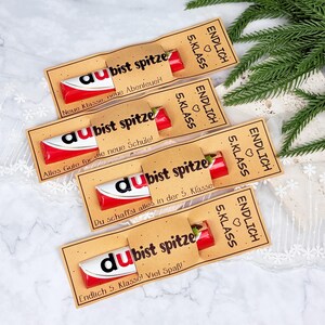 May include: Four chocolate bars wrapped in red and white packaging, each encased in a tan paper sleeve. The sleeves feature black text reading "ENDLICH 5. KLASS" and "du bist spitze," along with various German phrases like "Neue Klasse, neue Abenteuer!"