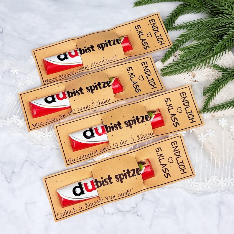 May include: Four chocolate bars wrapped in red and white packaging, each encased in a tan paper sleeve. The sleeves feature black text reading "ENDLICH 5. KLASS" and "du bist spitze," along with various German phrases like "Neue Klasse, neue Abenteuer!"