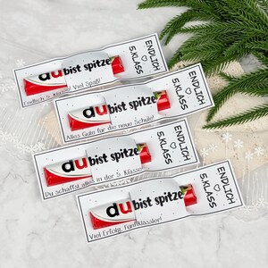 May include: Four red and white chocolate bars wrapped in white paper sleeves. Each sleeve features the black text 'du bist spitze' and 'ENDLICH 5. KLASS' with a small heart icon. The sleeves include various German phrases like 'Viel Spa&szlig;' and 'Viel Erfolg'.