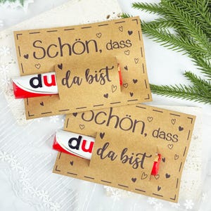 May include: Two kraft paper cards with the German text "Schön, dass du da bist" and heart illustrations. Each card holds a red and white candy bar. A sprig of greenery is in the background.