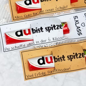 May include: Three chocolate bars wrapped in red and white packaging, each secured with a paper sleeve. The sleeves feature black text including 'du bist spitze', 'Alles Gute f&uuml;r die neue Schule', '5. KLASS', and 'Du schaffst alles in der 5. Klasse'.